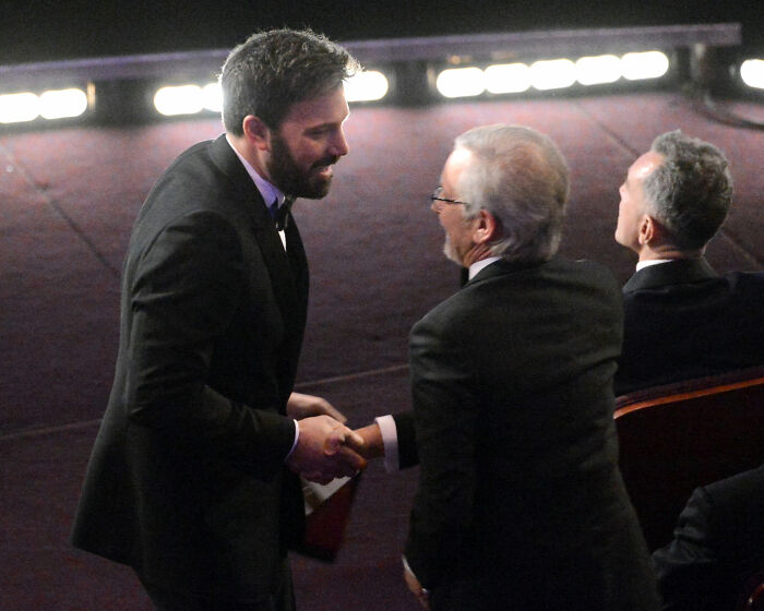 Ben Affleck shaking hands with Steven Spielberg at a formal event, both dressed in black suits and ties.