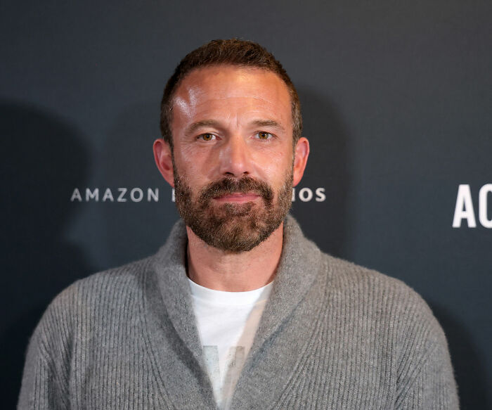 Ben Affleck at an Amazon Studios event, posing in a gray sweater with a trimmed beard and short hair.