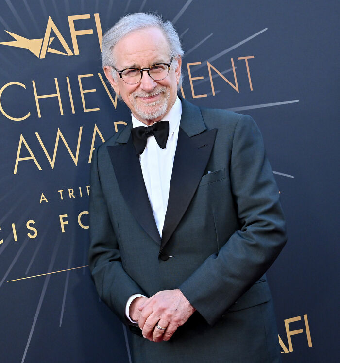 Steven Spielberg smiling at an awards event wearing a tuxedo with bow tie against a dark background.