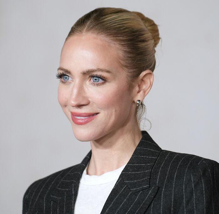 Britanny Snow smiling with hair in a bun, wearing a pinstripe blazer and white shirt against a plain background. Britanny Snow smiling with hair in a bun, wearing a pinstripe blazer and white shirt against a plain background.