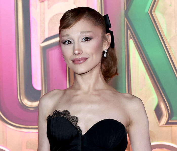 Ariana Grande posing in a black strapless dress with earrings, attending a colorful event background.