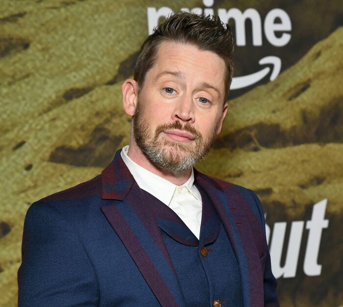 Actor Macaulay Culkin wearing a blue suit with burgundy accents, posing at a Home Alone related event.