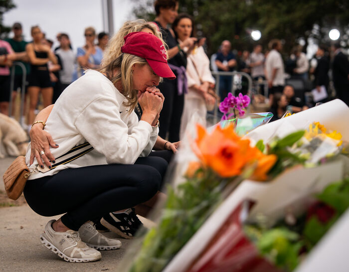 Woman in red cap mourns at a floral memorial as Bondi Beach attack and single mom's tone deaf GoFundMe spark outrage.