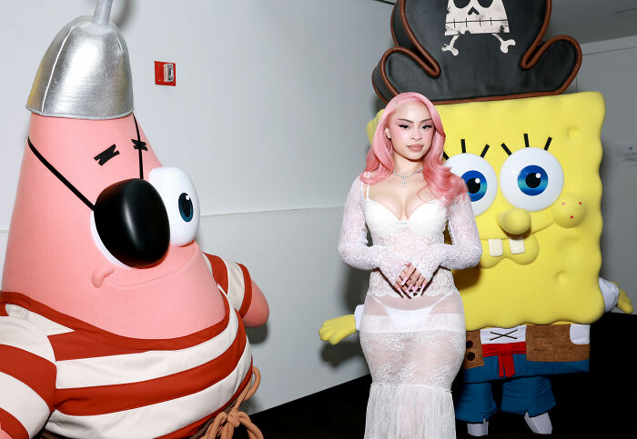 Ice Spice in a white see-through dress posing between Patrick Star and SpongeBob SquarePants characters at an event.
