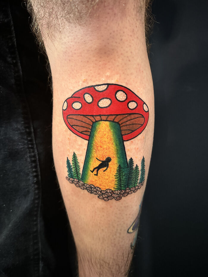 Colorful psychedelic tattoo on forearm features a red mushroom with a glowing green beam and tiny floating figure.