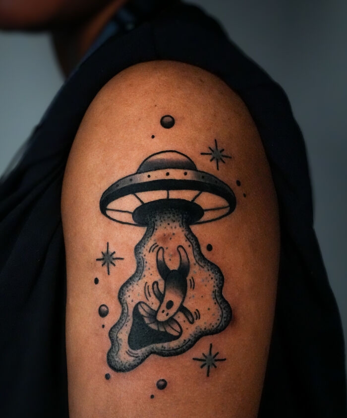 Black and gray psychedelic tattoo of a UFO abducting a creature surrounded by stars on a person's upper arm.