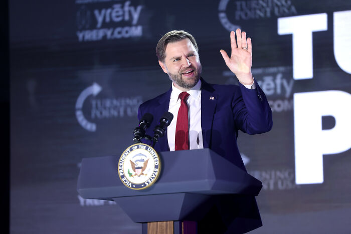JD Vance speaking at a podium with Vice President seal, addressing marriage speculation after Erika Kirk viral hug and Usha&rsquo;s bare finger.