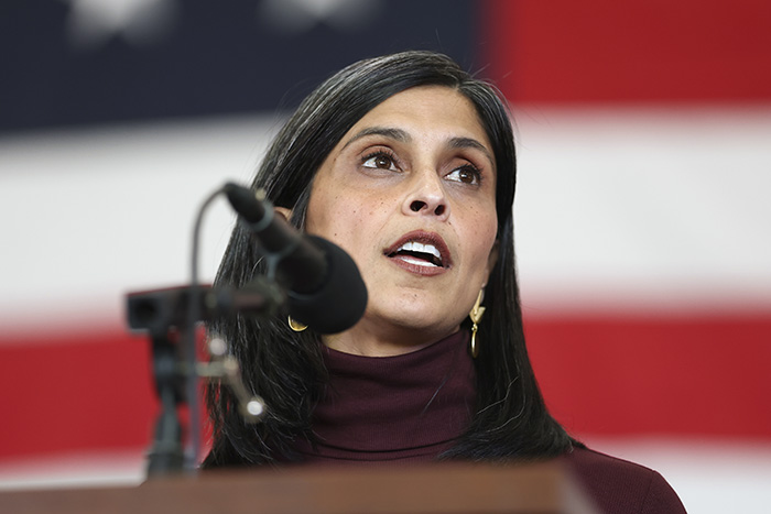 Usha Vance speaking at a podium with a microphone against an American flag background during a public event.