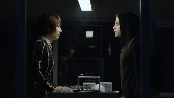 BD Wong from Jurassic World in a tense conversation wearing a striped outfit facing a person in a black hoodie indoors.