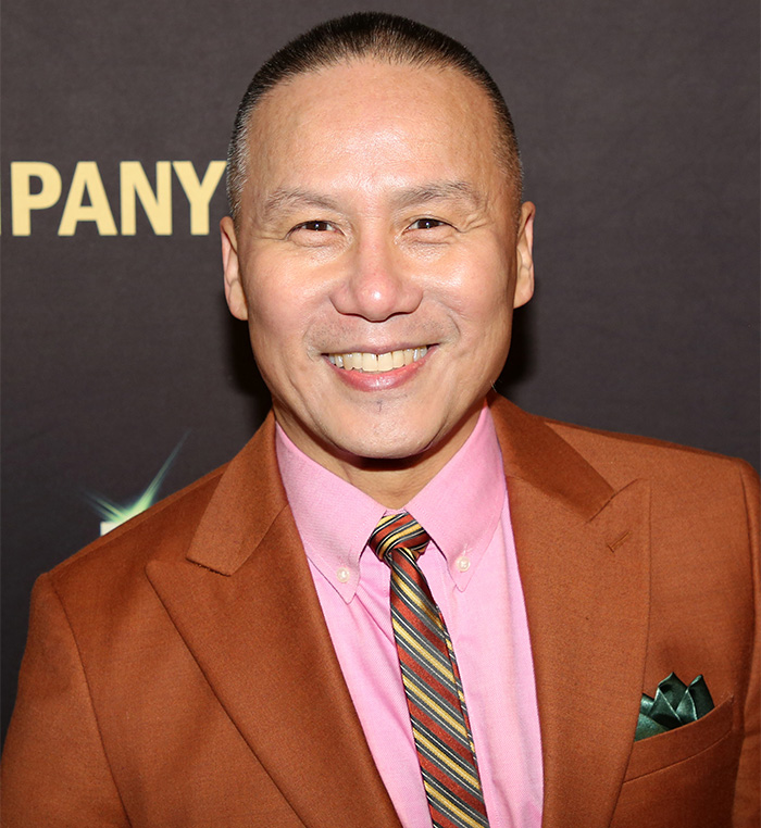 BD Wong, Jurassic World star, smiling in a brown suit and striped tie at a public event against a dark backdrop.