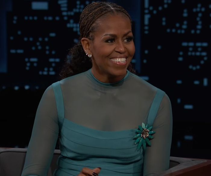 Michelle Obama smiling with bold makeup and braided hair during a TV interview praising makeup artist.