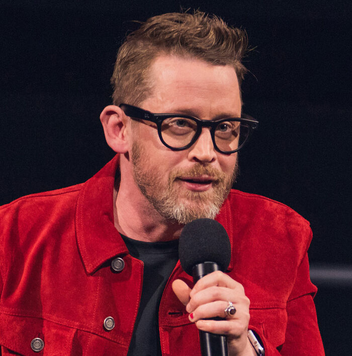 Macaulay Culkin speaking into a microphone, wearing glasses and a red jacket, addressing Home Alone Kevin&rsquo;s dad job theory.