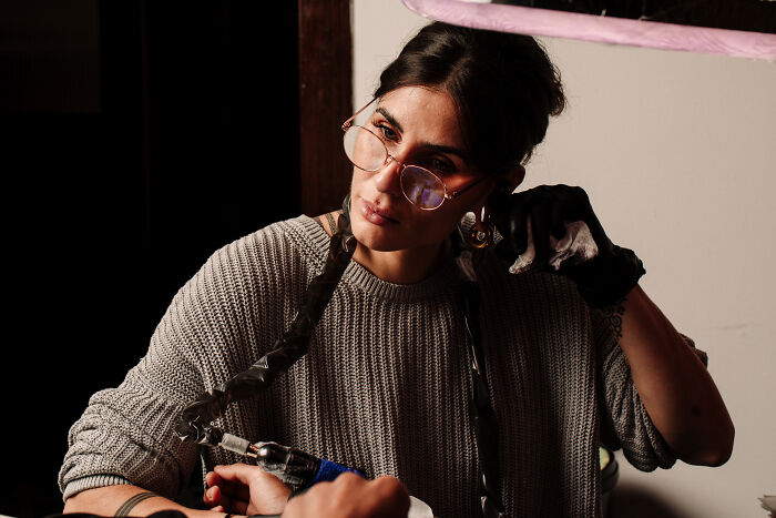 Young Sacred Gold artist with glasses and braids, focused on a creative tattoo session in a dimly lit studio.