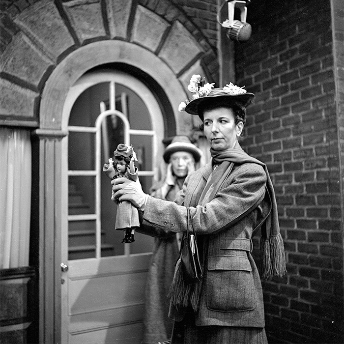 Vintage black and white rare pop culture shot of a woman holding a doll in period costume outside a brick building.
