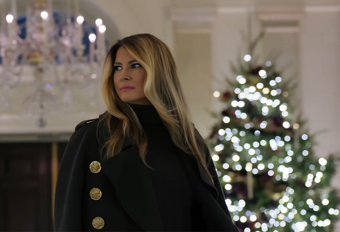 Melania Trump standing indoors near a decorated Christmas tree, related to her new Christmas ornament controversy.
