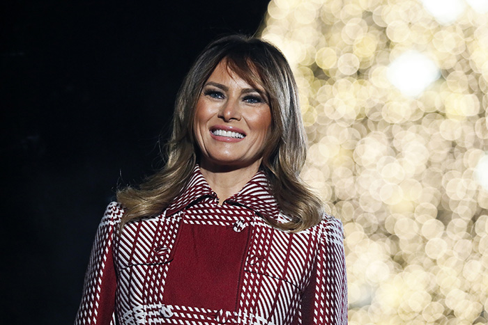 Melania Trump smiling in a red patterned coat with blurred warm lights in the background, linked to Christmas ornament outrage.