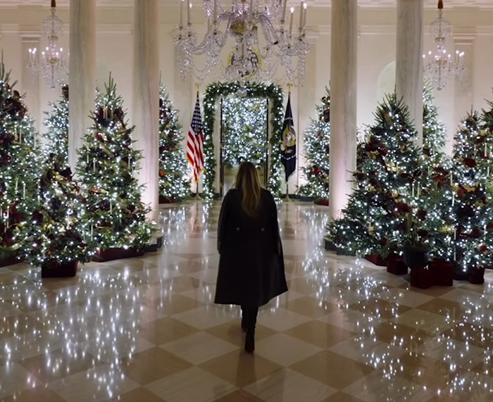 Melania Trump walking through a hall decorated with Christmas trees and sparkling lights for holiday season.