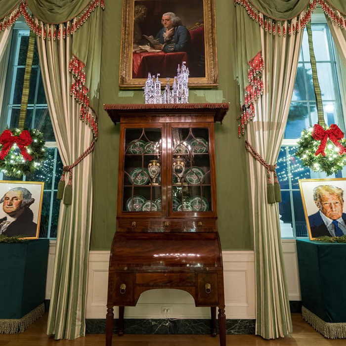 Christmas room display with Melania Trump's new Christmas ornament on wooden cabinet amid holiday wreaths and portrait paintings.