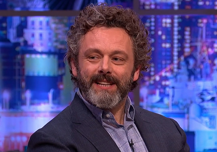 Michael Sheen smiling during a TV interview, wearing a suit jacket and checkered shirt in a colorful studio setting