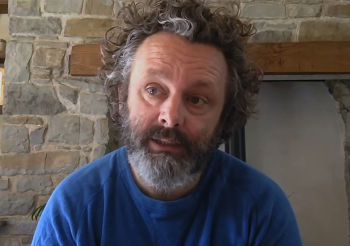 Michael Sheen with curly hair and beard, wearing a blue shirt, seated indoors near a stone wall and fireplace.