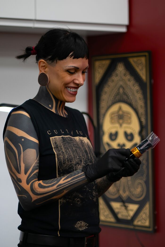 Sacred Gold artist with tattoos smiling while holding tattoo equipment in a studio with artwork in the background