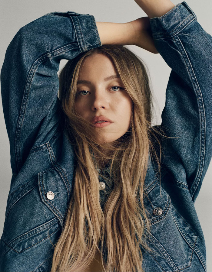 Sydney Sweeney posing in a denim jacket with arms raised, related to American Eagle ad apology and heated reactions.