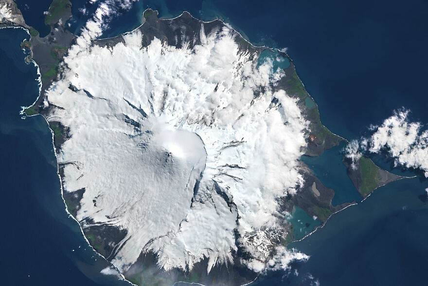Aerial view of a snow-covered volcanic island surrounded by ocean, representing dangerous places to visit in the world.