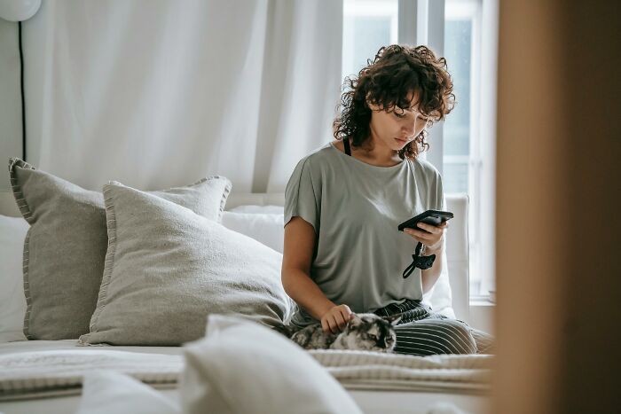 Woman sitting on bed calmly using phone while petting cat, illustrating staying calm in stressful scenarios.