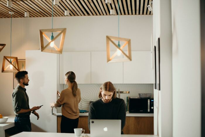 Three people in a modern kitchen with pendant lights, one using a laptop, others interacting, reflecting calm or tense scenarios.