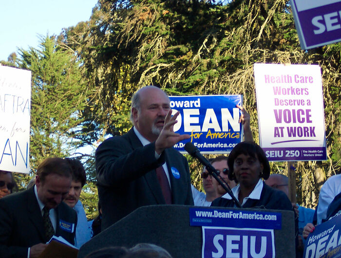 Man speaking at a political rally with SEIU signs, highlighting MAGA hypocrisy and comparisons between Rob Reiner and Charlie Kirk.
