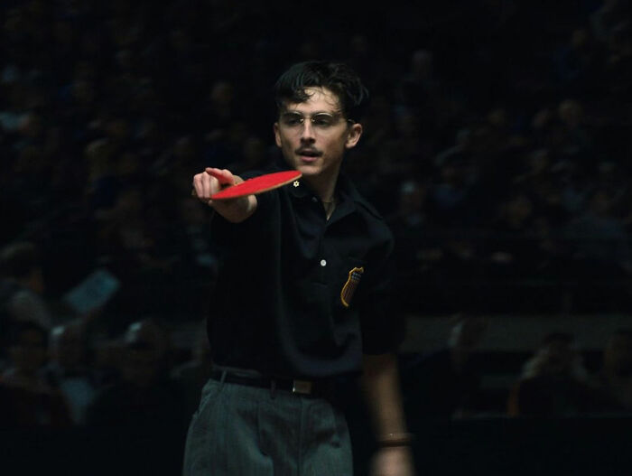 Young man in glasses playing table tennis, wearing a dark shirt and gray trousers in a dimly lit setting.