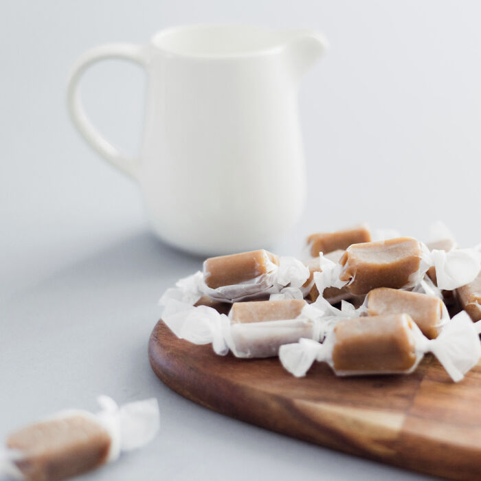 Caramel candies wrapped in white paper on a wooden board with a white ceramic pitcher in the background.