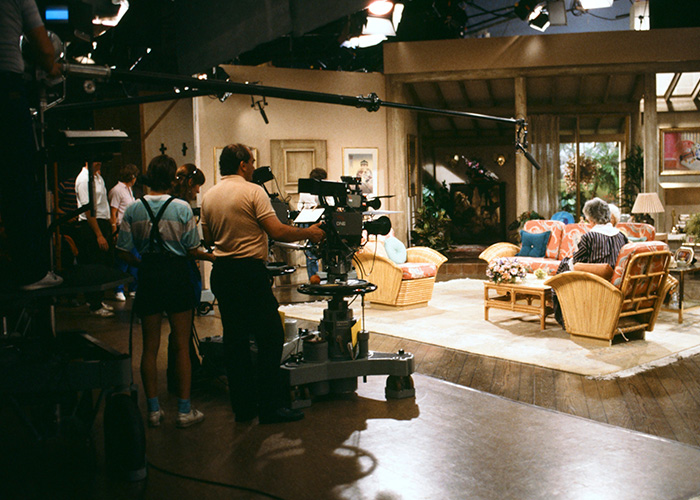 Behind-the-scenes pop culture shot of a TV show set with camera crew filming actors in a living room scene.