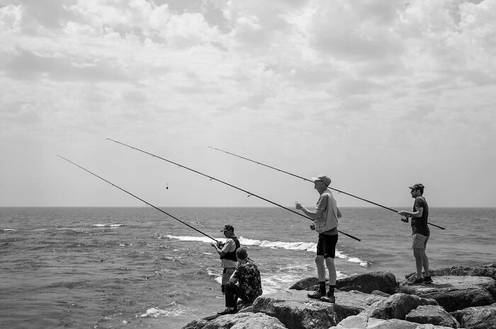 Four people fishing on rocky shore by the ocean, capturing marvels of daily life in street moments that feel like magic.
