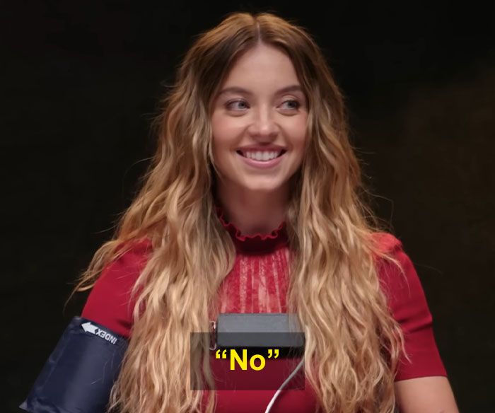Sydney Sweeney smiling during a lie detector test while wearing a red top and medical arm cuff in a dark room.