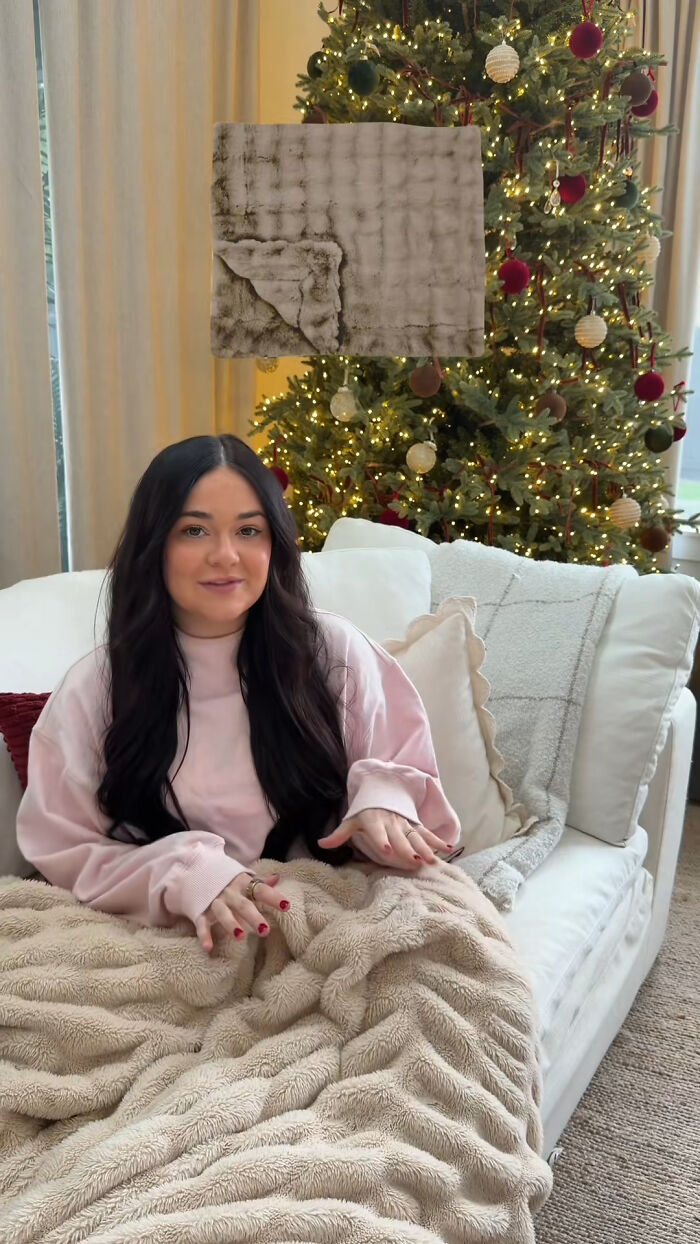 Woman sitting on cozy couch with plush beige blanket and decorated Christmas tree, showcasing prized Christmas gifts.