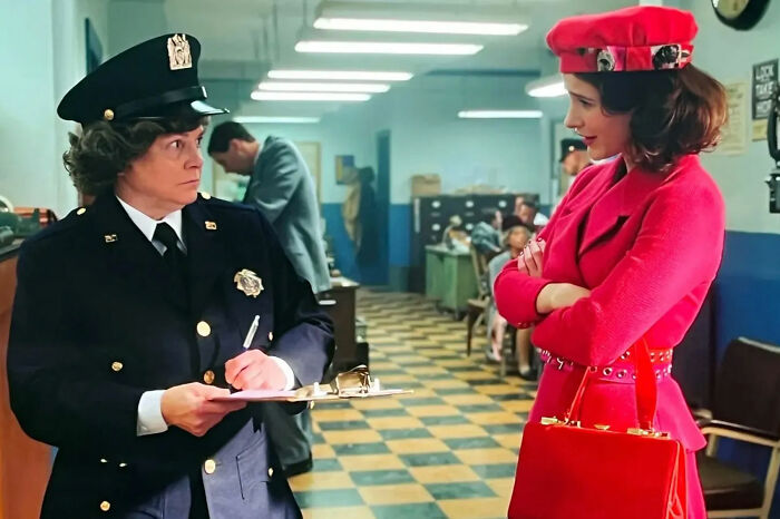 Actress from Marvelous Mrs. Maisel wearing red outfit standing in a police station talking to female officer with clipboard.