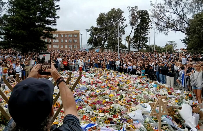 Large crowd gathers at a memorial filled with flowers, highlighting outrage after Bondi Beach attack and tone deaf GoFundMe.