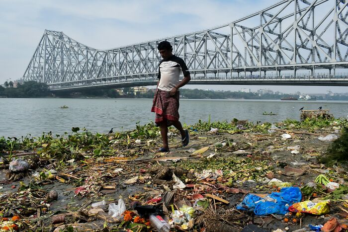Man walking on polluted riverbank with trash and plants near a large steel bridge, illustrating disturbing truths about pollution.