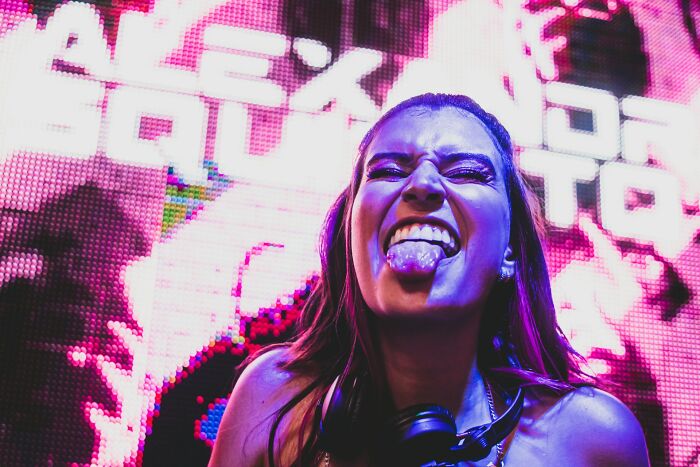 Young woman making a playful face with tongue out at a colorful neon-lit event, revealing unexpected dark secrets vibe.