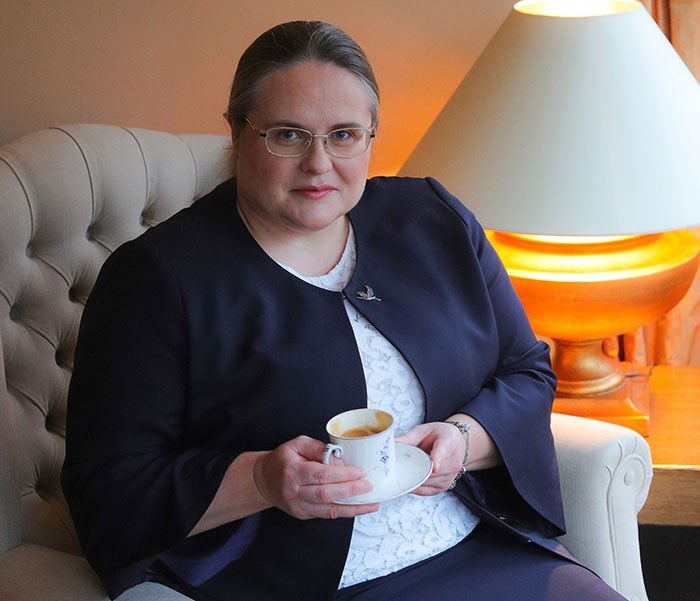 Woman in glasses holding a cup, seated indoors near a lamp, related to Lithuania lawmaker&rsquo;s cat consent news.