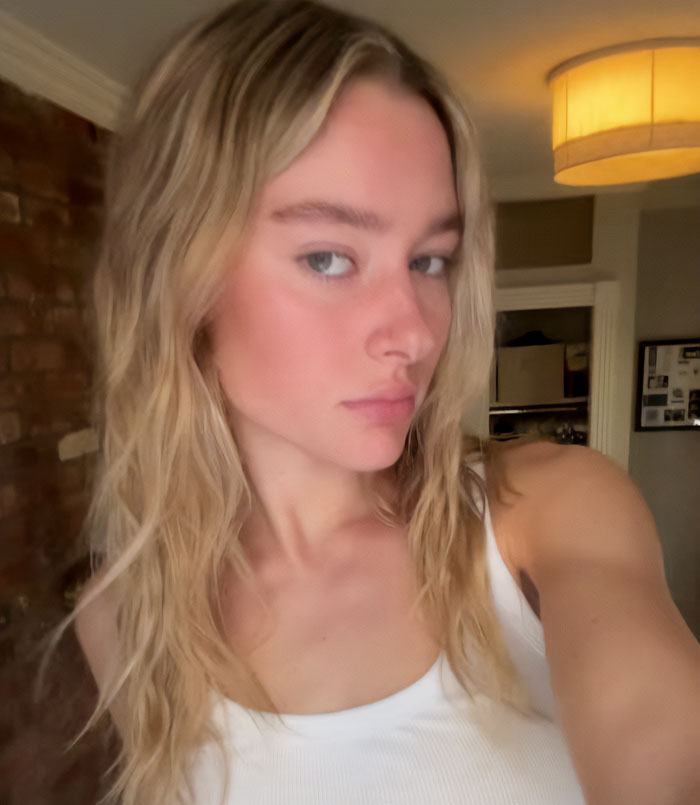 Young woman with long blonde hair wearing a white tank top inside a dimly lit room, related to NYU shove victim friend.