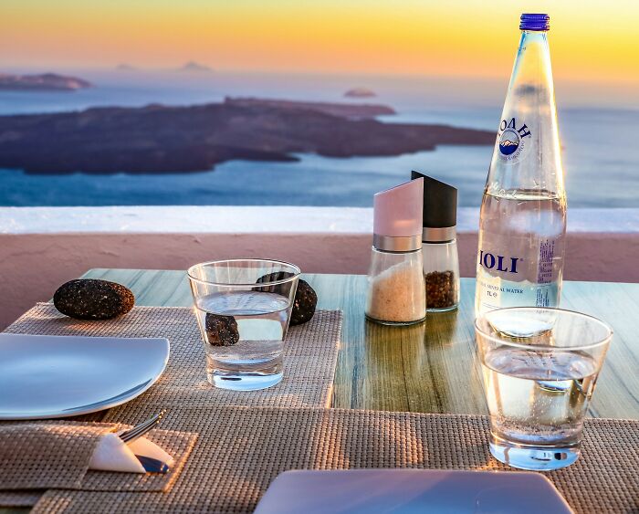 Table set with water glasses, salt, pepper, and bottle overlooking sea at sunset in a Greek village setting.