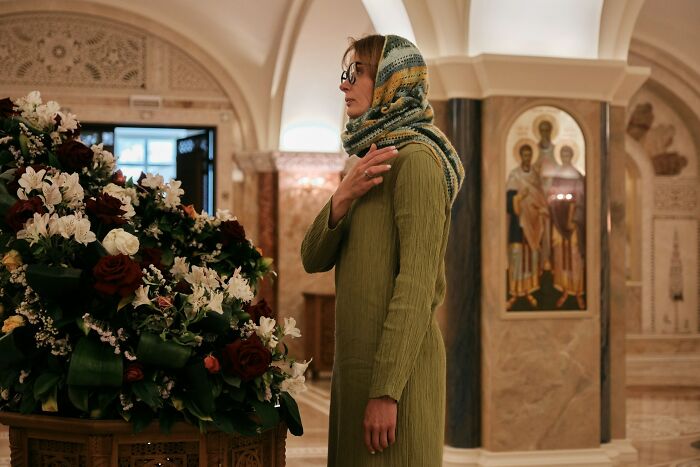 Woman in green dress and headscarf standing thoughtfully near floral arrangements inside an ornate funeral setting.