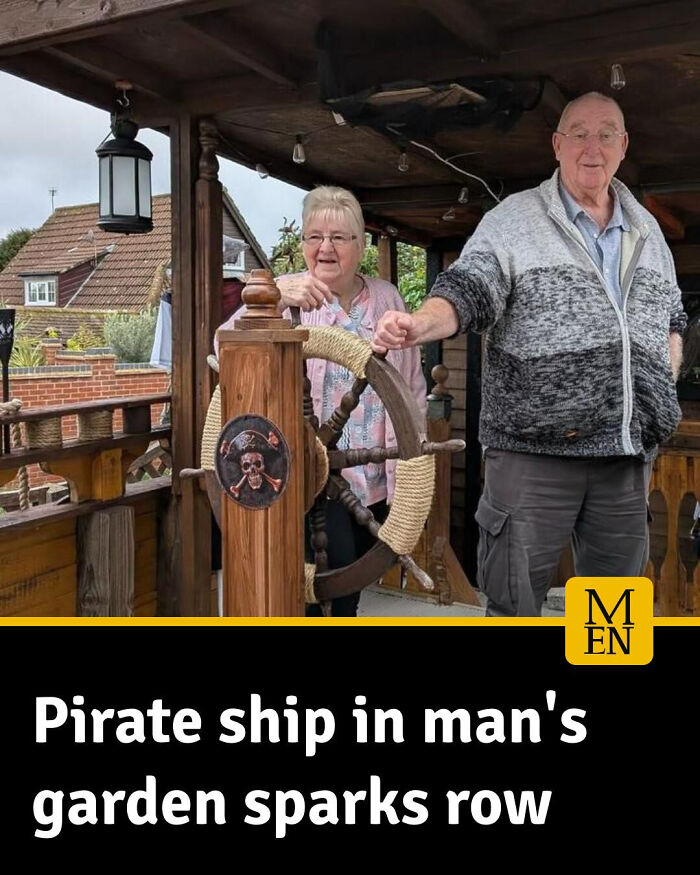 Two elderly people stand by a pirate ship wheel in a garden, a local newspaper covering unusual neighborhood events.