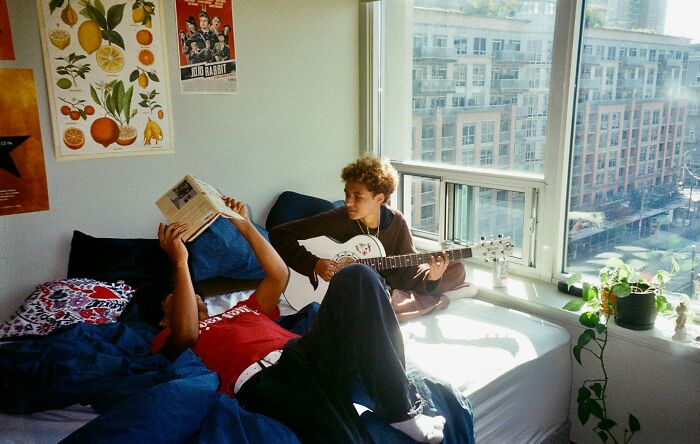 Two young people in a sunlit room, one reading a book and the other playing guitar, illustrating dark secrets revealed.