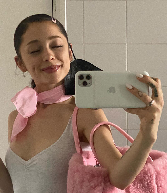 Ariana Grande taking a mirror selfie holding a phone, wearing a pink scarf and carrying a fluffy pink bag against a tiled background. Ariana Grande taking a mirror selfie holding a phone, wearing a pink scarf and carrying a fluffy pink bag against a tiled background.