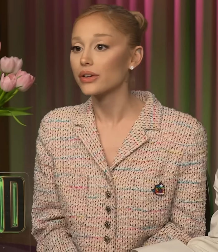 Ariana Grande speaking seriously about body-shaming, wearing a textured jacket with a floral arrangement nearby. Ariana Grande speaking seriously about body-shaming, wearing a textured jacket with a floral arrangement nearby.