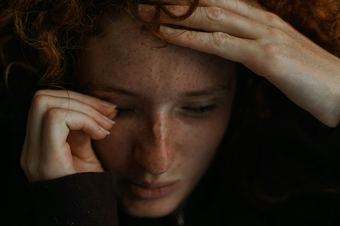 Young woman with red hair and freckles, holding her head in distress, portraying aunt pay niece tummy tuck drama emotions.