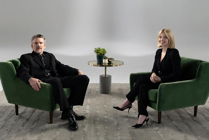 Ethan Hawke and Sydney Sweeney seated on green chairs in a minimalist studio setting, sparking online firestorm discussions.